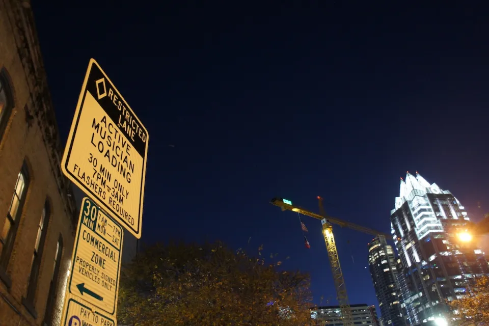 Close-up of musicians loading zone sign on Sixth Street in Austin, Texas at night with illuminated high-rise with pyramidal top in background.